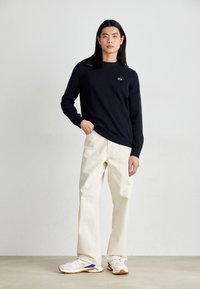 Chemise en coton bleu marine à manches longues avec col rond, logo brodé ; associée à un pantalon large beige et des baskets blanches avec des accents bleus.