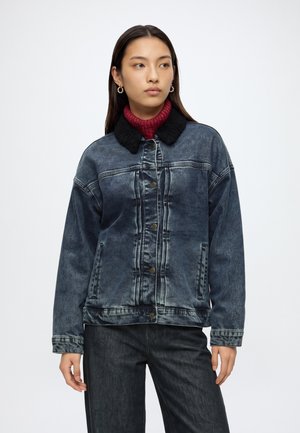 Jeune femme portant une veste en jean foncé délavée avec un col noir et un pull à col roulé rouge, regardant légèrement sur le côté.