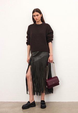 Donna che indossa un maglione marrone scuro, una gonna in pelle nera con frange e spacco alto, scarpe loafers nere chunky, mentre tiene una borsa borgogna con catena dorata.