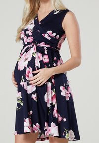 Gravid kvinna i ärmliv klänning i marinblått med rosa blommönster, händerna varsamt omfamnar hennes mage mot en enkel bakgrund.