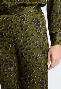 Olijfgroene broek met een zwart en bruin onregelmatig luipaardpatroon, gedragen met een bijpassend topje, hand ontspannen aan de zijkant.