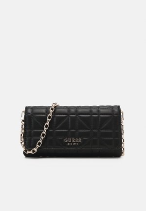 Pochette matelassée noire Guess avec bandoulière chaîne argentée et logo Guess à l'avant, sur fond clair uni.