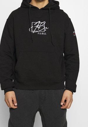 Mężczyzna w czarnej bluzie z kapturem z kieszenią z przodu i białym logo F.U.B.U. na środku klatki piersiowej, w połączeniu z ciemnymi spodniami.