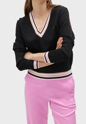 Personne portant un pull noir en V avec des rayures roses, blanches et dorées, les bras croisés, et un pantalon rose sur un fond uni.