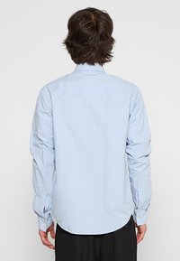 Camicia azzurra chiara con maniche lunghe, cucitura sulla spalla sul retro e polsini con un solo bottone. Il tessuto appare liscio e leggero.