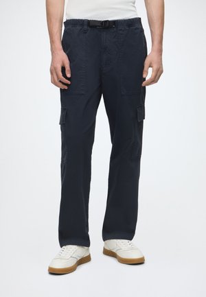 Mann trägt schwarze Cargohosen mit mehreren Taschen, einen schwarzen Gürtel, weiße Sneaker und ein sichtbarer Teil eines weißen Hemdes.