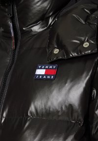 Chaqueta acolchada negra con un acabado brillante, presenta secciones acolchadas horizontales, un cierre de cremallera y un parche con el logo "TOMMY JEANS" en el pecho.
