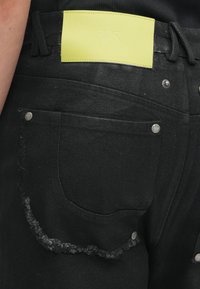 Crne traperice s izrezanim rubom na džepu, s svijetložutim kožnim etiketom na pojasu s utisnutim tekstom.