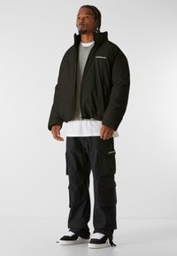 Chaqueta de plumas negra con cuello alto, logo blanco en el pecho, combinada con un suéter de punto gris, pantalones cargo negros y zapatillas negras.