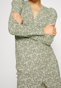 Grünes Blumenkleid mit langen Ärmeln, V-Ausschnitt und gerafftem Detail, ausgestattet mit einem kleinen weißen Blumenmuster und einer glatten Stofftextur.