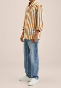 Camicia a righe in senape e bianco, con colletto a abottatura e maniche arrotolate, abbinata a jeans azzurri larghi e sneakers bianche.
