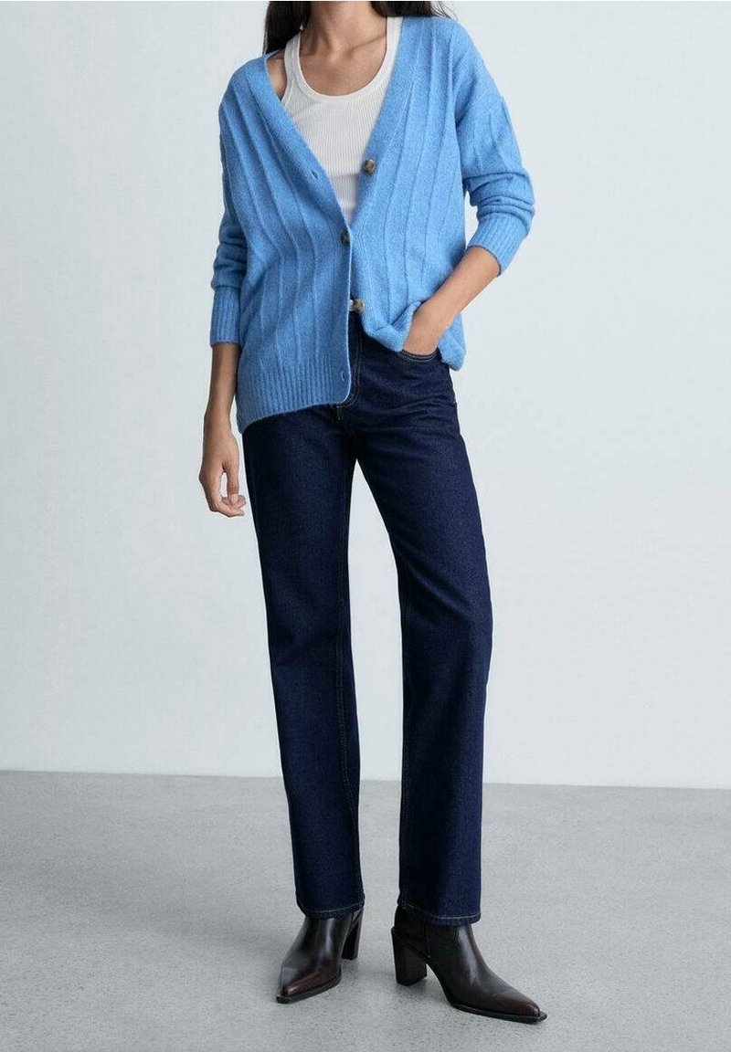 Femme debout portant un cardigan bleu clair boutonné, un débardeur blanc, un jean bleu foncé et des bottines noires à talons sur un sol gris.