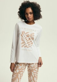 Biała koszula z długim rękawem w roślinnym nadruku w jasnobrązowym kolorze, z tekstem. Uzupełniona beżowymi spodniami w liściasty wzór.