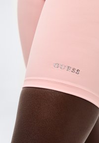 Κοντινό πλάνο σκούρου μηρού που φοράει ανοιχτό ροζ σορτς με ασημένιο λογότυπο "GUESS" κοντά στο τελείωμα.