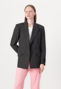 Dvojradový blazer v tmavosivej farbe s bielymi pásikmi, so zúženými klopami a čiernymi gombíkmi; skombinované so svetloružovými nohavicami.