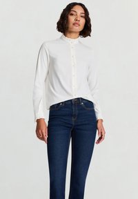 Weiße Bluse mit langen Ärmeln, einem gerafften Kragen und Knopfleiste vorne, kombiniert mit dunkelblauen Jeans mit geradem Bein. Glatter Stoff, minimale Details.