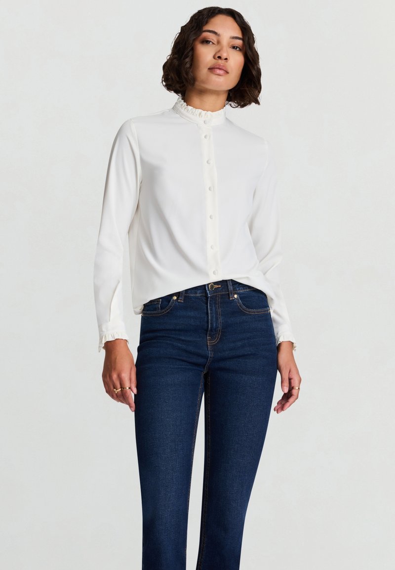 Weiße Bluse mit langen Ärmeln, einem gerafften Kragen und Knopfleiste vorne, kombiniert mit dunkelblauen Jeans mit geradem Bein. Glatter Stoff, minimale Details.
