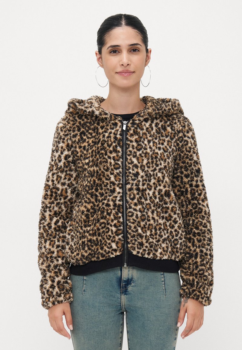 Leopardmønstret hettegenser med glidelås i myk, teksturert, brun og svart kunstpels, med hette og ribbestrikkede mansjetter og kant. Kombinert med lyseblå jeans.