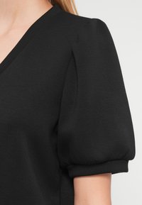 Gros plan d'une personne portant un haut noir à manches ballon avec un col en V et des poignets froncés sur des manches courtes.