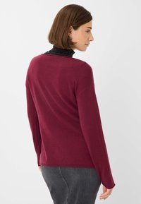Maglione bordeaux con maniche lunghe, scollo a V e una consistenza a maglia morbida. Indossato sopra una camicia nera con colletto, visto da dietro.