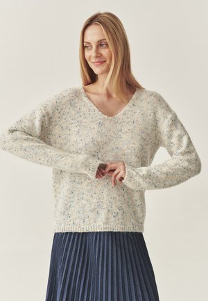 Hellbeige gestrickter Pullover mit mehrfarbigen Sprenkeln, V-Ausschnitt, überschnittenen Schultern und gerippten Bündchen, kombiniert mit einem gefalteten dunklen Rock.