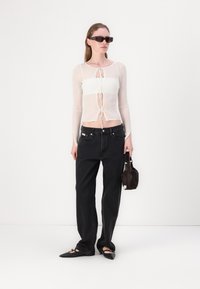 Langermet, gjennomsiktig hvit topp med rynket design, sammen med løse svarte jeans og spisse svarte leiligheter, holder en liten brun veske.