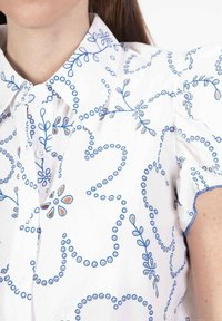 Chemise blanche à boutons avec broderie florale bleue et accents circulaires, manches courtes bouffantes et col.