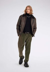 Braune Lederjacke mit einem schwarzen Kunstfellkragen, getragen über einem schwarzen Rollkragenpullover, kombiniert mit olivgrünen Cordhosen und schwarzen Schuhen.