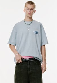 Ung mand iført en lyseblå oversized T-shirt med teksten "STWD GOALS", mørke bukser, naglet bælte og lag-dekoreret halskæde mod hvid baggrund.