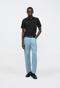 Schwarzes Poloshirt, hellblaue Jeans mit geradem Bein, schwarzer Gürtel und schwarze Schuhe. Der Stoff wirkt weich, mit einem klassischen Kragen.