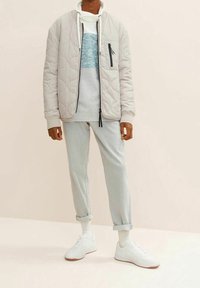 Giacca bomber beige trapuntata, maglione grigio con striscia blu, pantaloni grigi chiari a risvolto e sneaker bianche. Design minimalista con accenti funzionali.