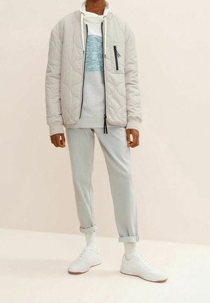 Vadderad beige bomberjacka, grå tröja med blå rand, ljusgrå uppvikta byxor och vita sneakers. Minimalistisk design med funktionella detaljer.