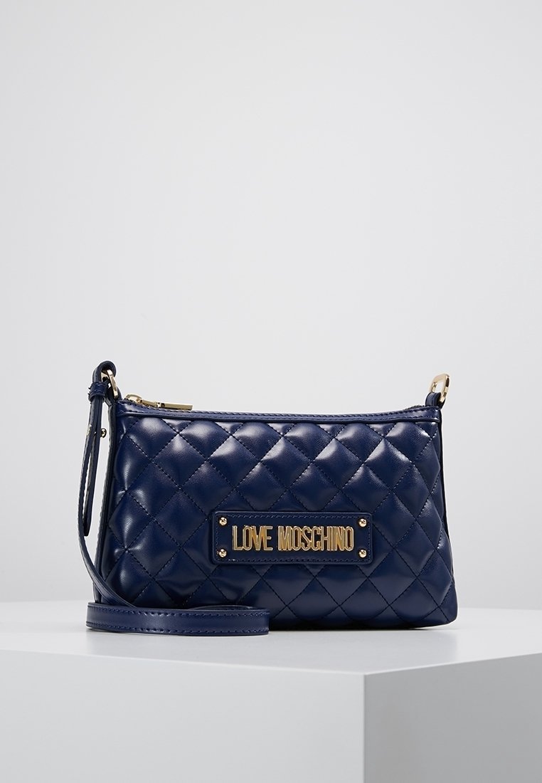 Prošívaná tmavě modrá kožená crossbody taška se zlatým kováním a obdélníkovou značkovou destičkou. Obsahuje odnímatelný popruh a zipové zavírání.