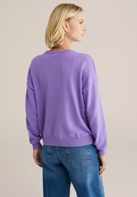 Purpurový sveter s uvoľneným strihom, pletenou textúrou, spustenými ramenami, ribbovaným lemom a manžetami, kombinovaný s modrými džínsami s vysokým pásom.