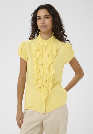 Žena v žltej blúzke s volánikmi a nadýchanými rukávmi a béžových textúrovaných nohaviciach, stojaca s jednou rukou na páse proti jednoliatemu pozadiu.