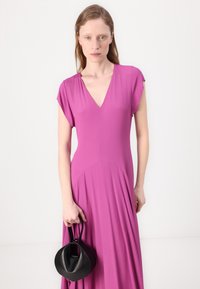 Puku V-neck, mikono fupi, iliyotengenezwa kwa kitambaa laini na kinachoelea kwa rangi ya pinki angavu, na muonekano wa mtiririko. Mfano anashikilia mkoba mduara mweusi.