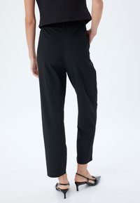 Schwarze maßgeschneiderte Hose mit geradem Bein, ausgestattet mit einer Gesäßtasche und aus glattem Stoff. Kombiniert mit eleganten schwarzen Absatzschuhen.