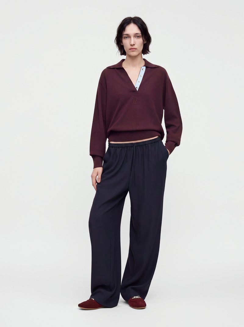 Woman standing wearing a maroon long-sleeve shirt with blue embroidered detail and loose black pants with maroon slip-on shoes.