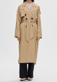 Person i en lång beige trenchcoat med midjebälte, svarta byxor, ljusa skor och en grå överdel mot en neutral bakgrund.