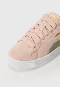 Baskets en daim rose avec une texture douce, dotées de lacets plats, d'une semelle en caoutchouc blanche et d'un accent vert sur le côté. Étiquette logo dorée sur la languette.