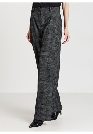 Femme portant un pantalon large à carreaux gris taille haute et des bottes à talons noires à bout pointu, debout devant un fond uni.