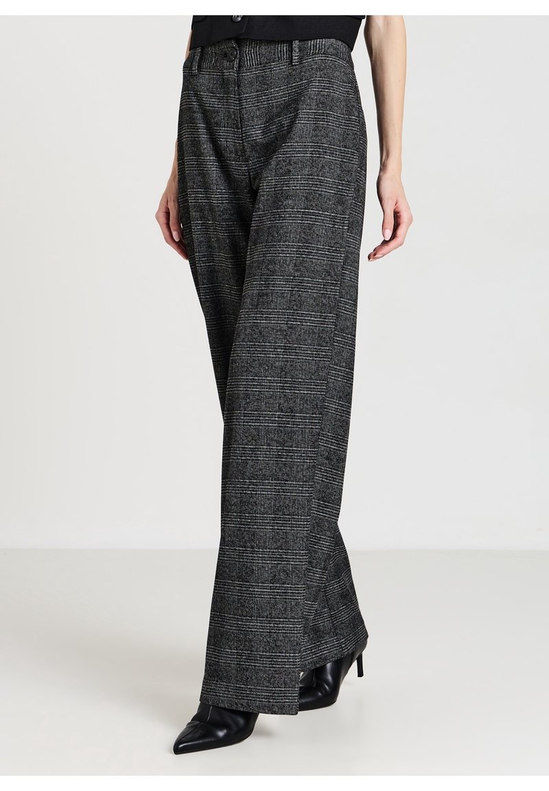 Femme portant un pantalon large à carreaux gris taille haute et des bottes à talons noires à bout pointu, debout devant un fond uni.