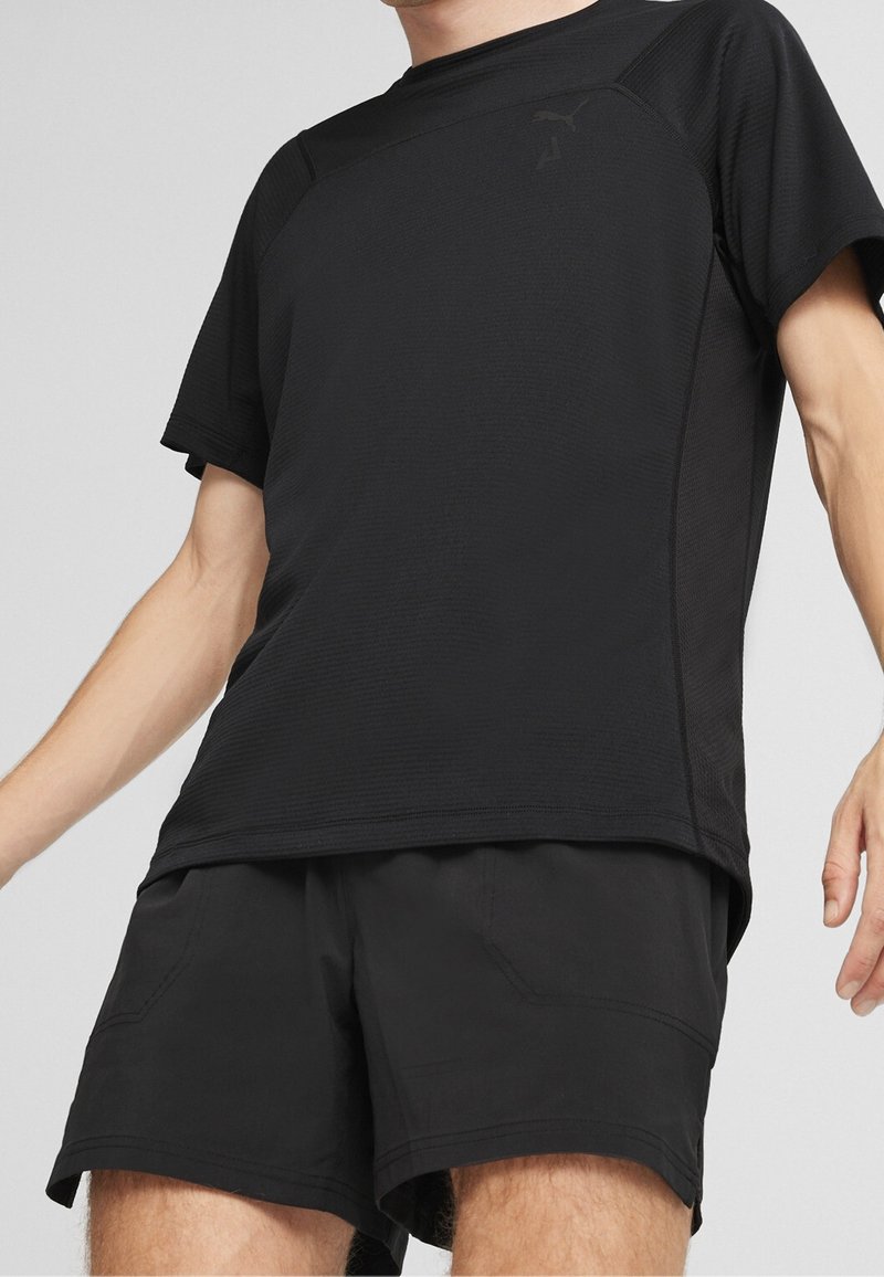 Personne portant un t-shirt de sport noir à manches courtes et un short noir, visible de la poitrine aux cuisses sur un fond uni.