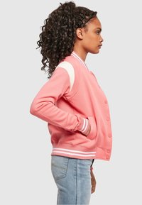 Roza varsity jakna s bijelim detaljima, rebrastim manšetama i kopčanjem na kopče. Mekani materijal i opušteni kroj, stilizirana s svijetloplavim trapericama.