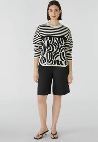 Gebreide trui met zwart-witte strepen en abstracte patronen, gecombineerd met zwarte shorts en zwarte sandalen met straps. Soepele textuur.