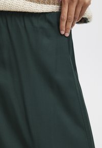 Jupe vert foncé avec une taille élastique, en tissu lisse et mat. La couture latérale est subtilement visible, ce qui renforce la structure du vêtement.
