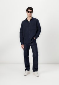Navy button-up shirt with a single chest pocket, paired with dark denim jeans and white sneakers in front of a plain background.