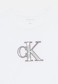 Balta medvilninė marškinėlių su dideliu sidabriniu "ck" logotipu, apibrėžtu rožine spalva. Klasikinis apvalus iškirpimas ir trumpomis rankovėmis.