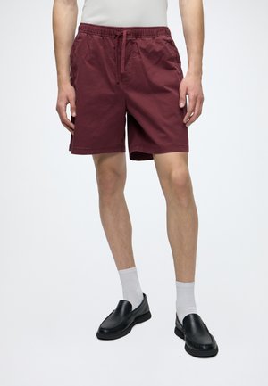 Uomo che indossa pantaloncini bordeaux fino al ginocchio con coulisse, calzini bianchi alti e scarpe nere slip-on, in piedi davanti a uno sfondo semplice.