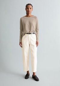 Frau mit geflochtenem Haar, die ein braun-weiß gestreiftes Langarmshirt, cremefarbene Hosen mit einem schwarzen Gürtel und dunkelbraune Loafers trägt und steht.